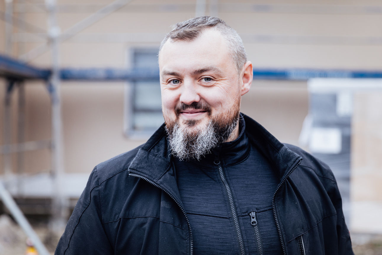 Friedrich Wilhelm befindet sich auf einer aktiven Baustelle eines Wohnbauprojekts der  „Friedrich Bau“. Er ist umgeben von Bauelementen und Gerüststrukturen, die den aktuellen Baufortschritt und das Arbeitsumfeld im Wohnungsbau widerspiegeln