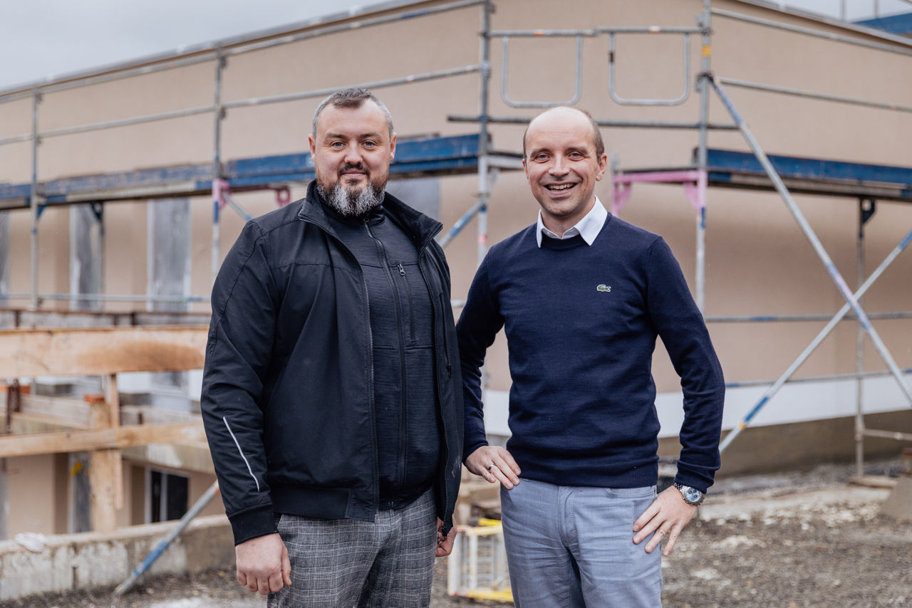 Wilhelm Friedrich und Berater Marco Müller befinden sich auf der Baustelle eines Neubauprojekts der „Friedrich Bau“. Der eingerüstete Gebäudebereich zeigt den aktuellen Stand der Bauarbeiten und dokumentiert die laufende Baufortschrittskontrolle im Rahmen des Projekts