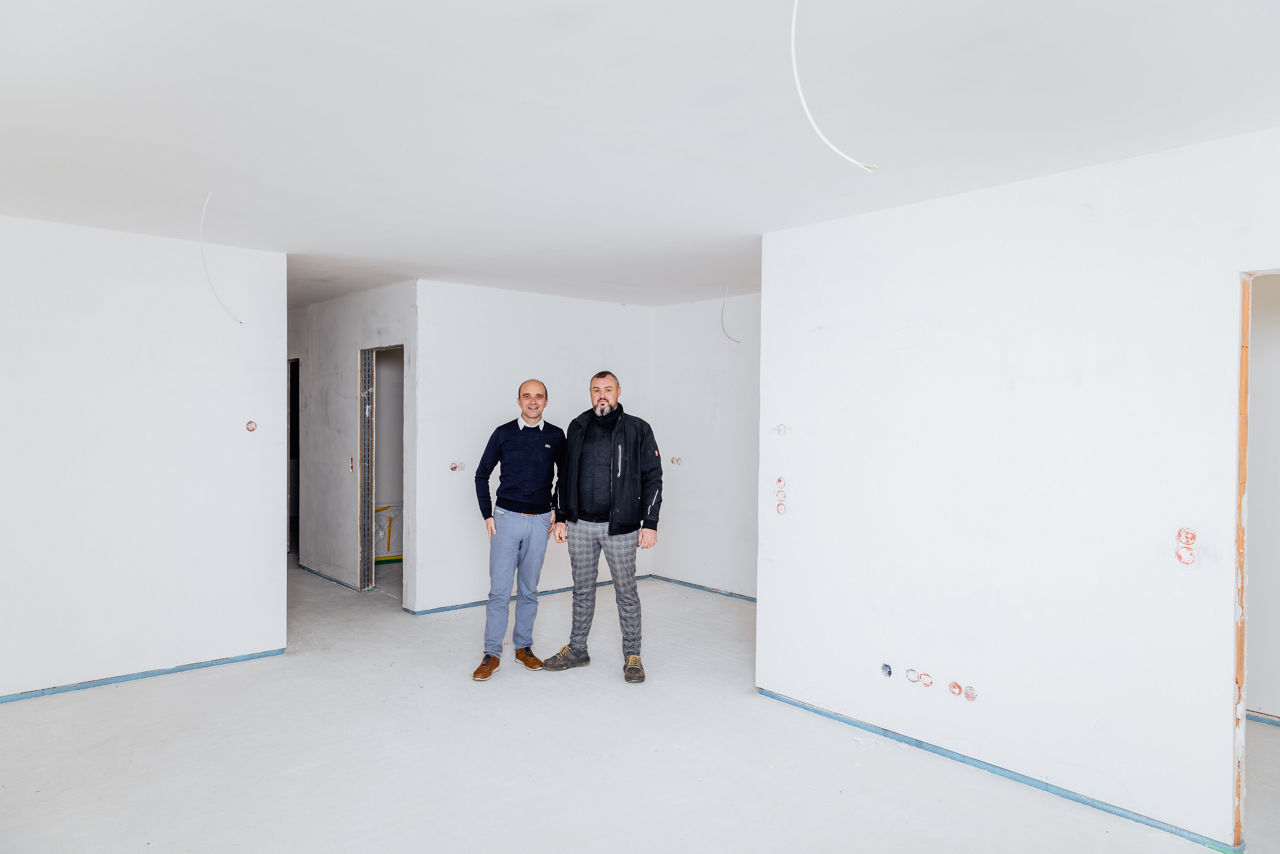Wilhelm Friedrich und Berater Marco Müller befinden sich in einem weitgehend fertiggestellten Rohbau eines Neubauprojekts von „Friedrich Bau“. Die Aufnahme zeigt einen zukünftigen Wohnbereich mit sichtbaren Baustrukturen und dokumentiert den aktuellen Stand der Ausbauarbeiten im regionalen Neubauprojekt