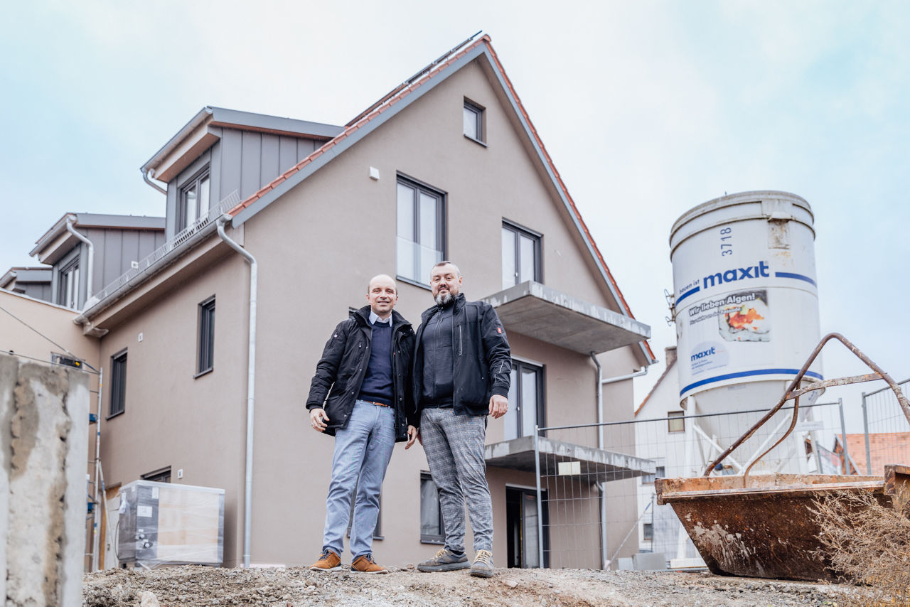 Berater Marco Müller und Wilhelm Friedrich stehen vor einem mehrgeschossigen Neubauprojekt von „Friedrich Bau“. Die Baustellenszene zeigt sichtbare Infrastruktur und Baugeräte und dokumentiert den aktuellen Stand laufender Wohnbauprojekte in der Region Main‑Rhön