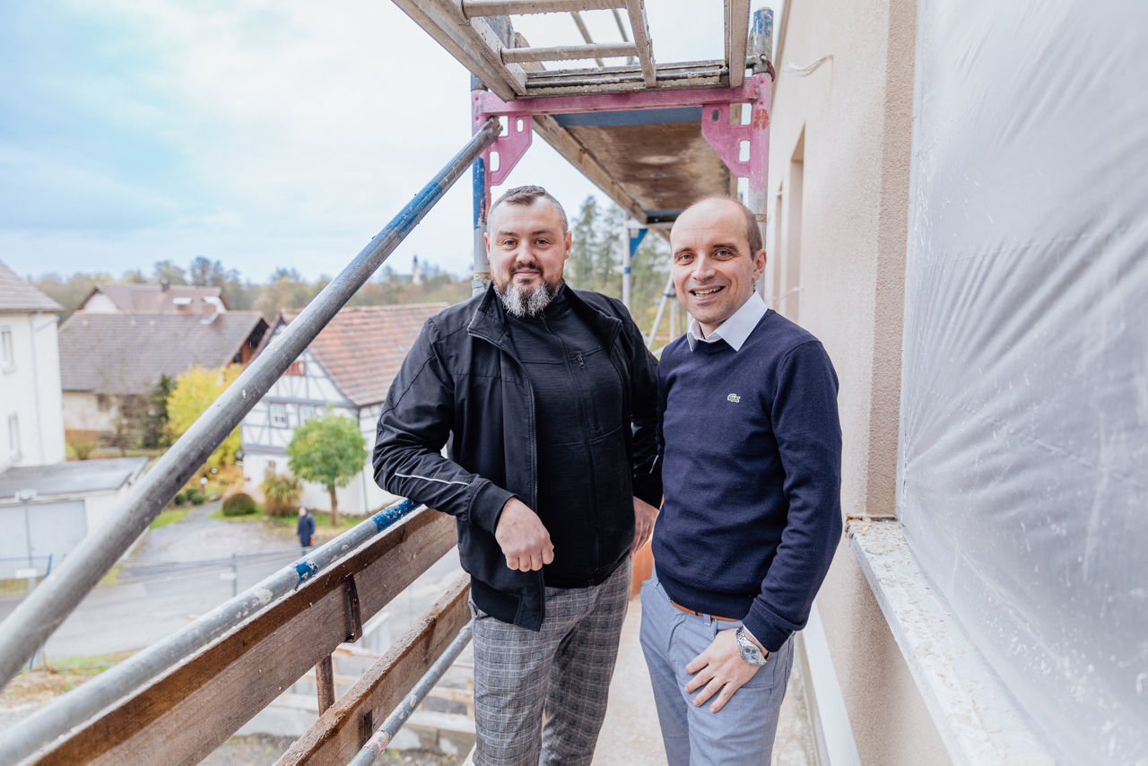 Wilhelm Friedrich und Berater Marco Müller befinden sich auf dem Außengerüst eines mehrgeschossigen Wohnbauprojekts von „Friedrich Bau“ in der Region Main‑Rhön. Die Aufnahme zeigt den Baustellenbesuch während des Baufortschritts mit Blick auf Gerüstkonstruktionen und die äußere Gebäudestruktur