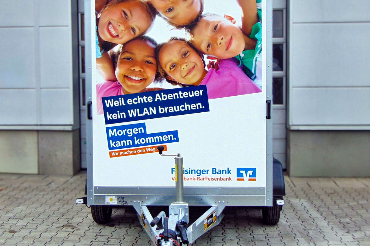 Frontalfoto eines Kfz-Anhängers mit Werbefläche der Freisinger Bank: Foto von 5 Kindern die im Kreis nach unten in die Kamera schauen; darunter Schriftzug: Weil echte Abenteuer kein WLAN brauchen 