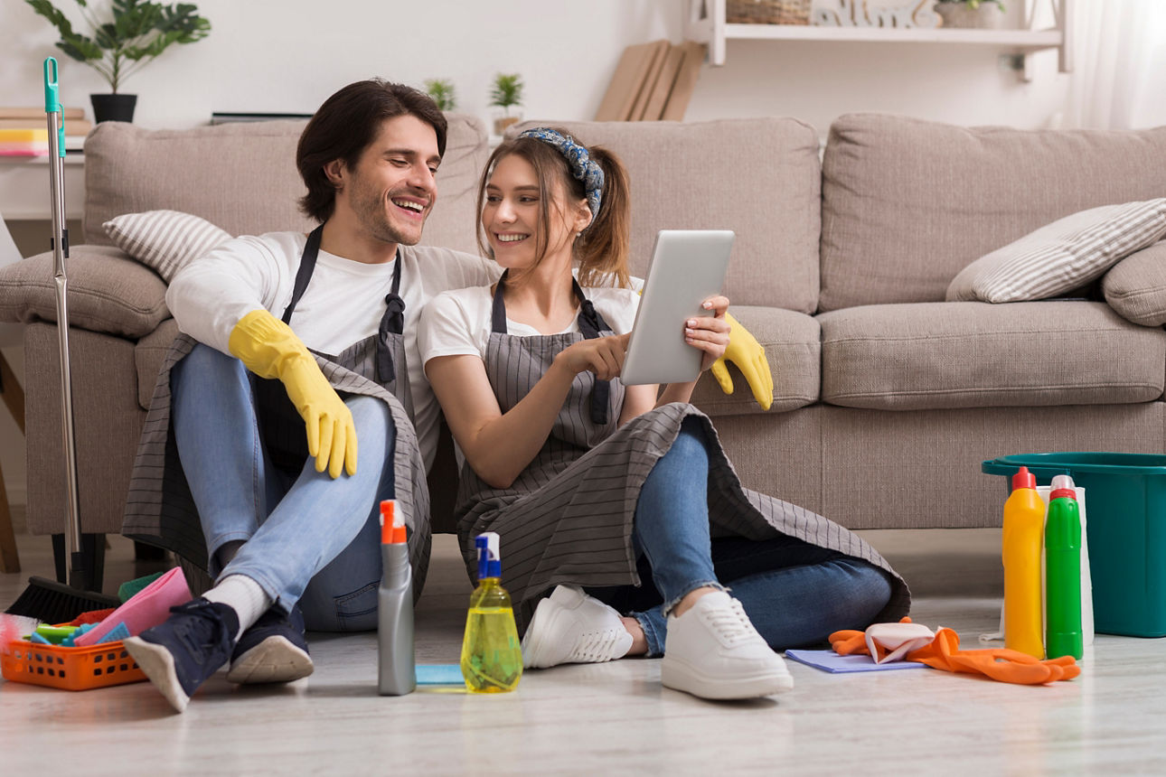 Fröhliches junges Paar, das sich nach dem Putzen der Wohnung mit einem Tablet entspannt und online Essen bestellt oder im Internet surft.