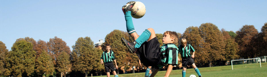 Ein Kind in Fußballkleidung führt auf einem Spielfeld einen Fallrückzieher aus, während Mitspieler zuschauen.