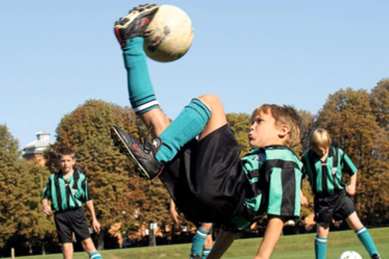 Ein Kind in Fußballkleidung führt auf einem Spielfeld einen Fallrückzieher aus, während Mitspieler zuschauen.