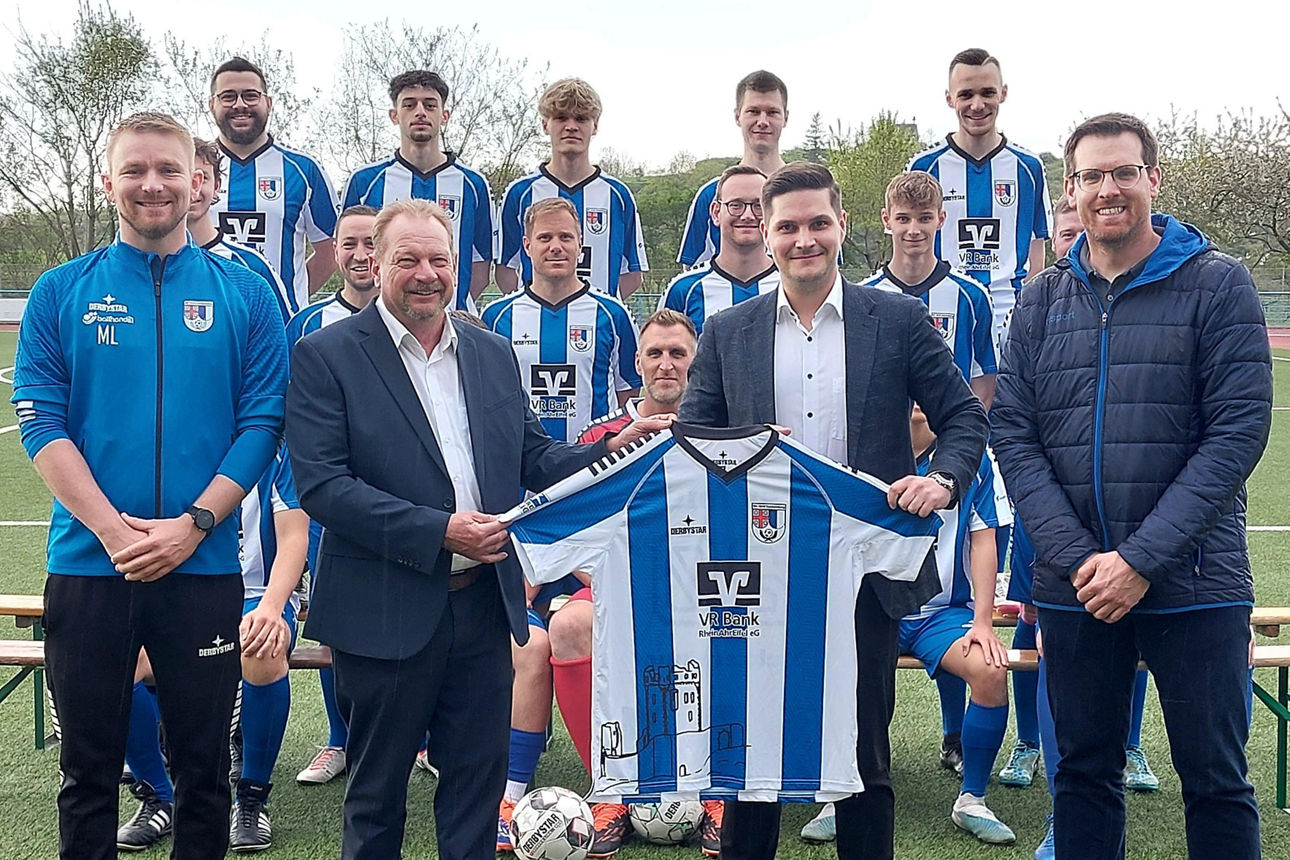 Fußballspieler stehen auf dem Fußballplatz zusammen mit Funktionären und Bankvertretern in einem Gruppenbild zusammen.