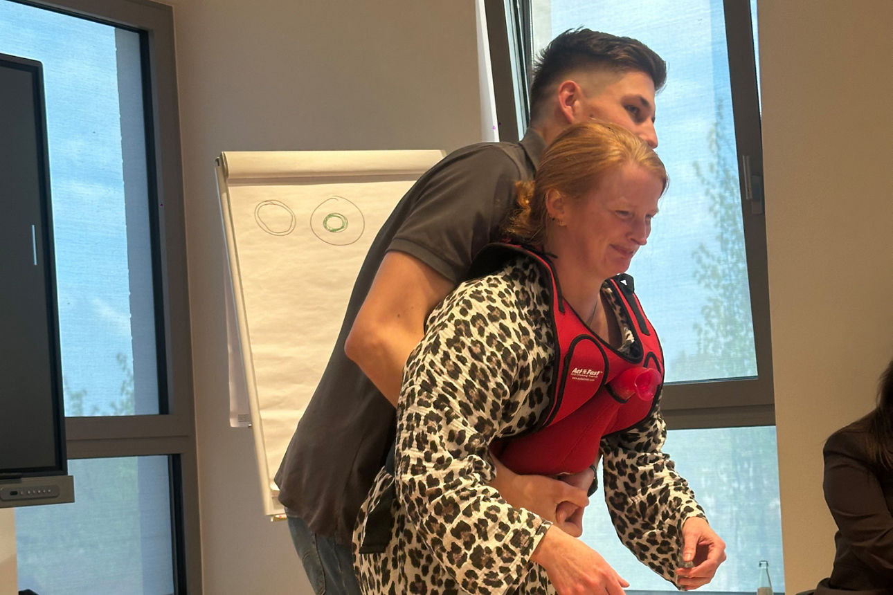 Frauen und Finanzen: Erste Hilfe Übung - Ein Mann steht hinter einer Frau und umfasst ihren Bauchraum.