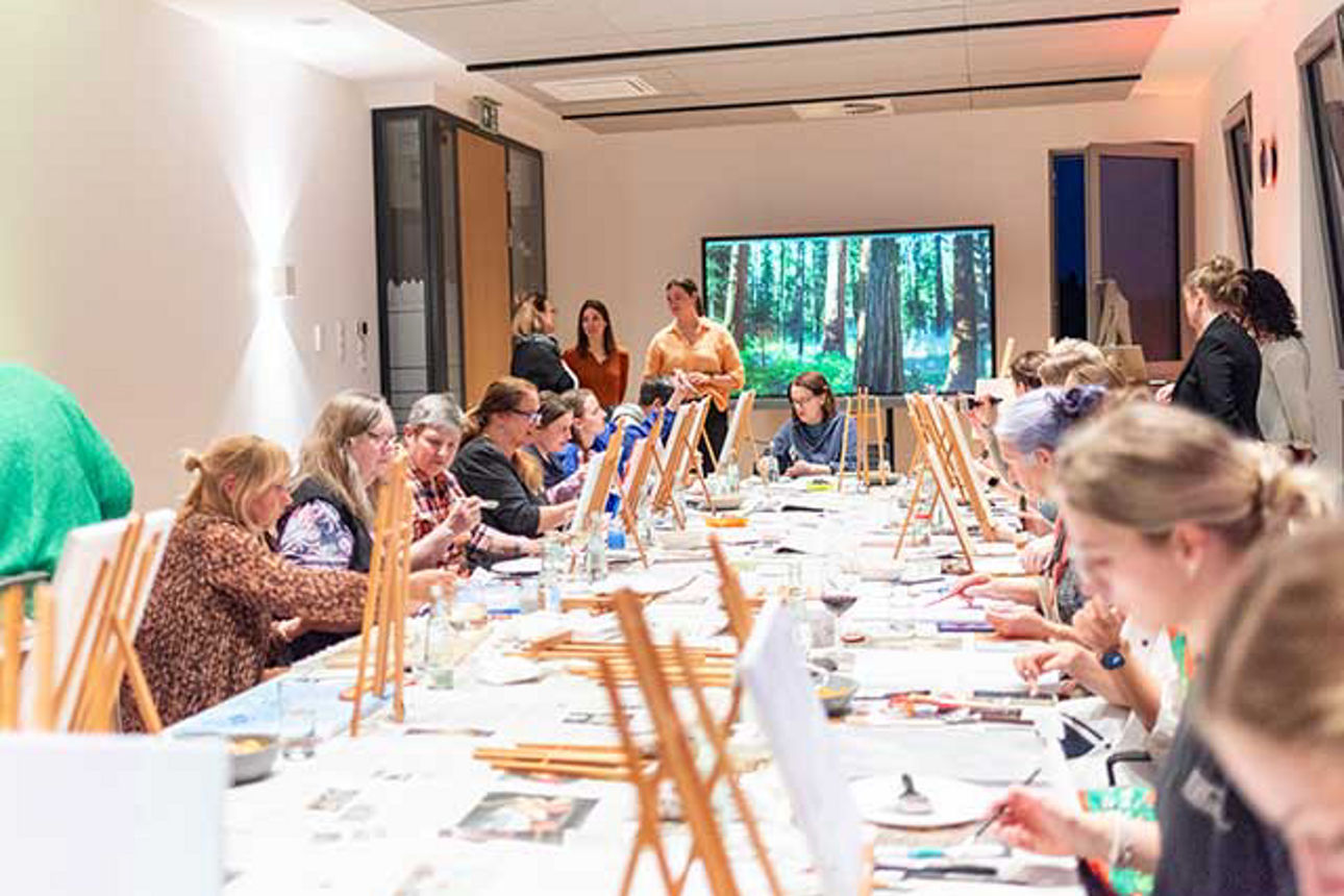 Frauen sitzen an einem großen Tisch und malen auf kleinen Staffeleien.