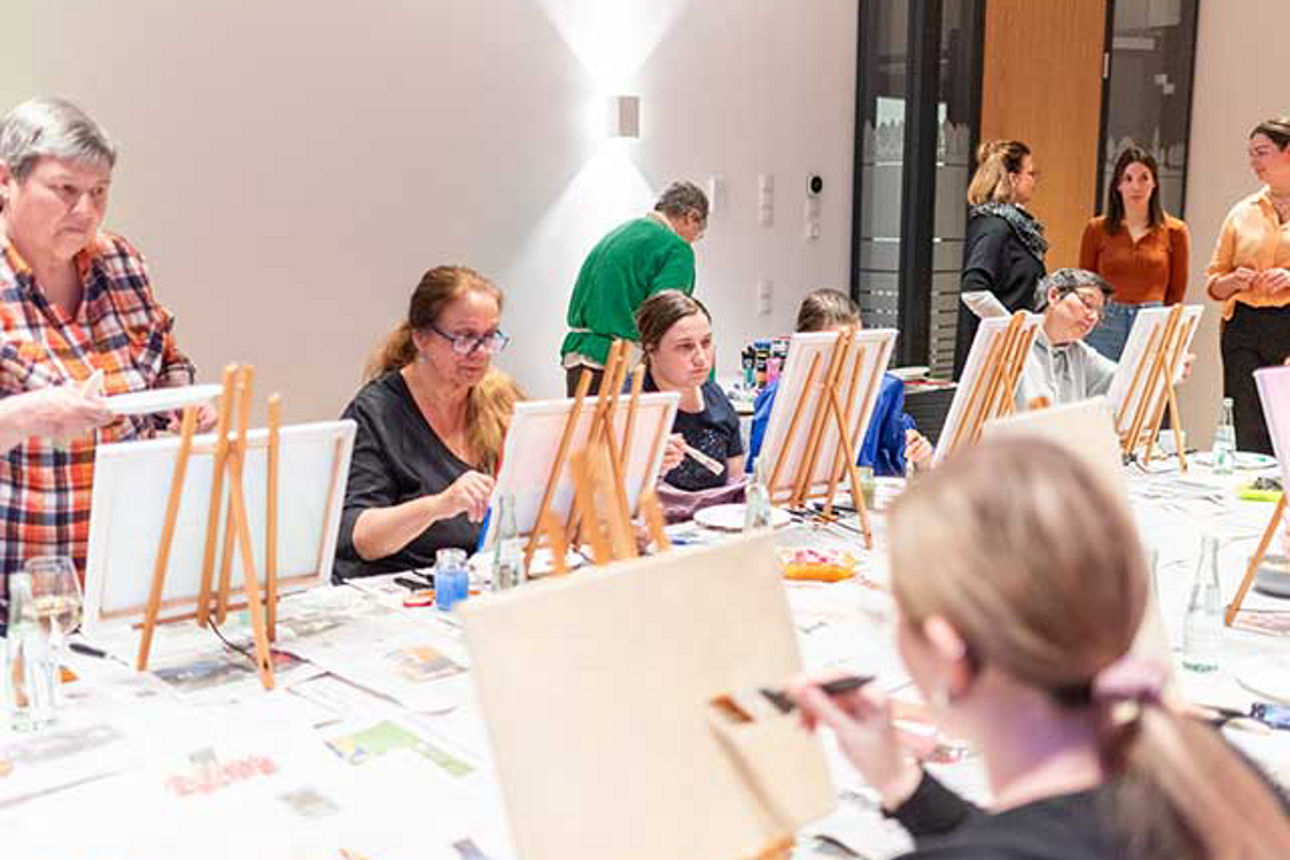 Frauen sitzen an einem großen Tisch und malen auf kleinen Staffeleien.