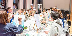 Frauen sitzen an einem langen Tisch vor einer Staffelei und malen auf Leinwand.