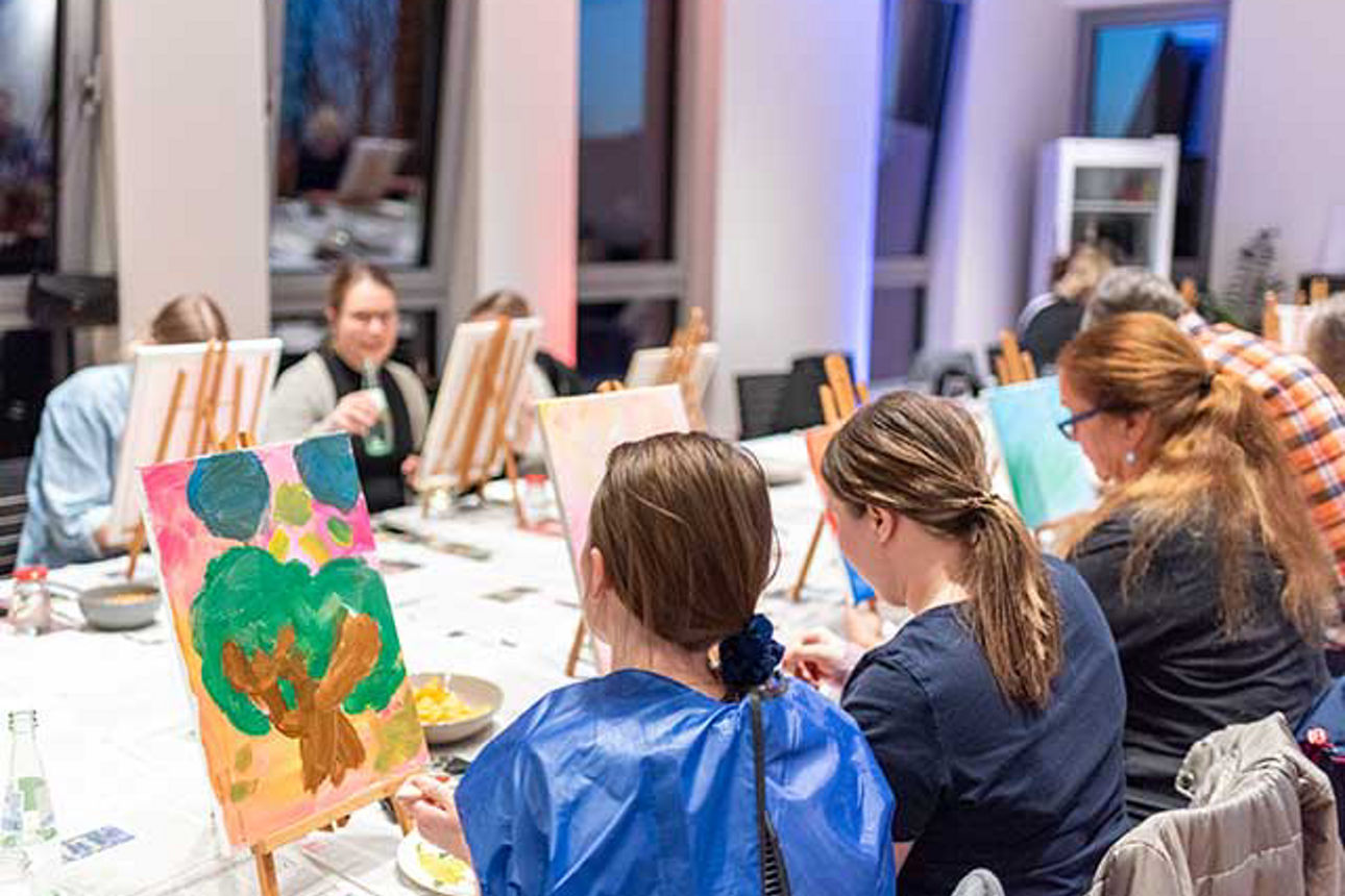 Frauen sitzen an einem großen Tisch und malen auf kleinen Staffeleien. Auf einem Bild ist ein Baum gemalt.