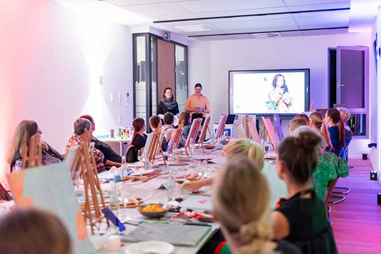  An einem langen Tisch sitzen Frauen mit ihren Staffeleien und folgen einem Vortrag.