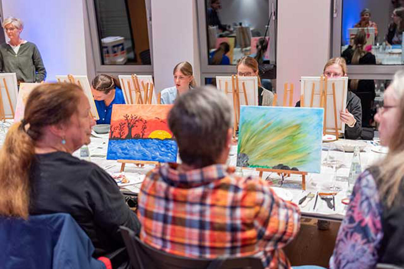  An einem langen Tisch sitzen Frauen mit ihren Staffeleien. 
