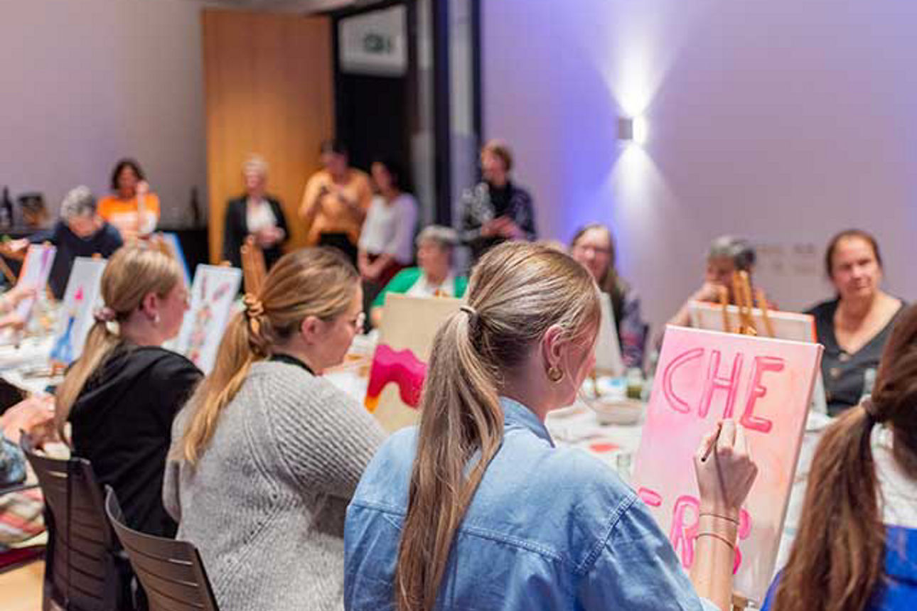  An einem langen Tisch sitzen Frauen mit ihren Staffeleien. 