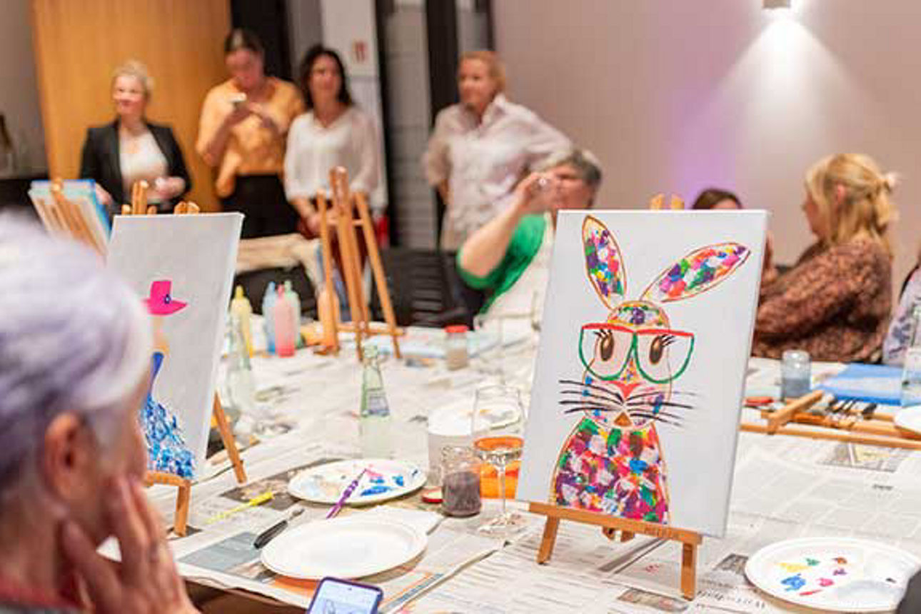  An einem langen Tisch sitzen Frauen mit ihren Staffeleien. Auf einem Bild wurde ein Osterhase gezeichnet.