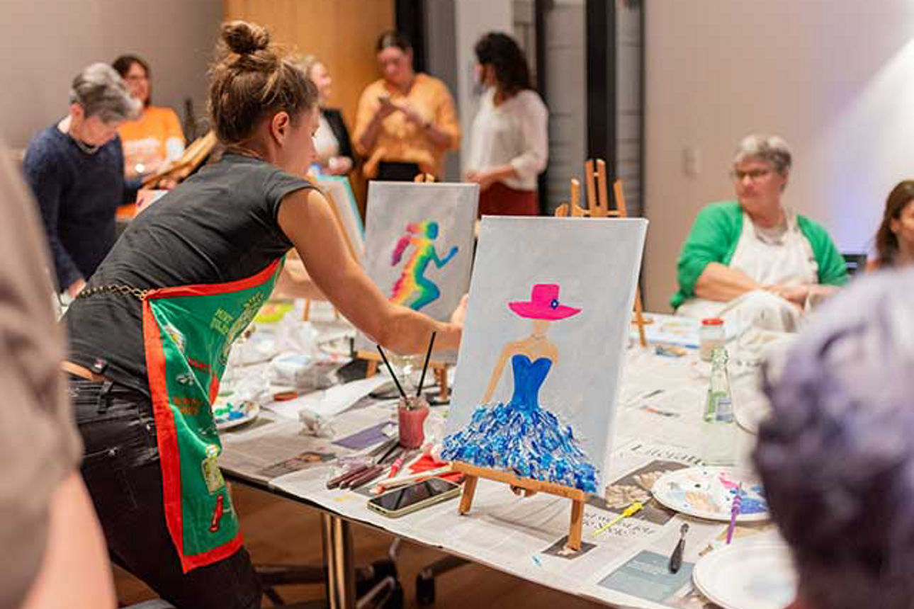  An einem langen Tisch sitzen Frauen mit ihren Staffeleien. 