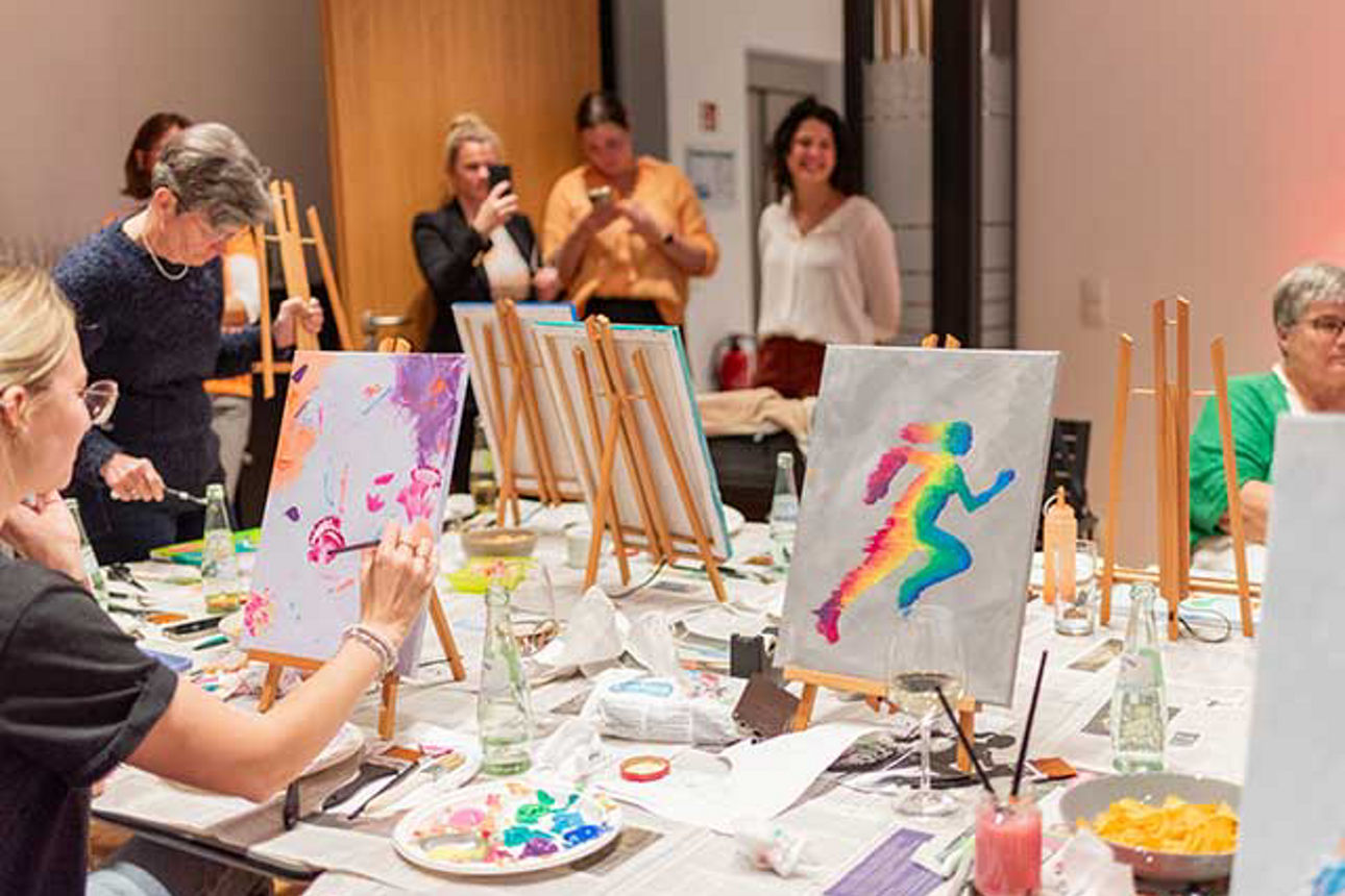  An einem langen Tisch sitzen Frauen mit ihren Staffeleien. 