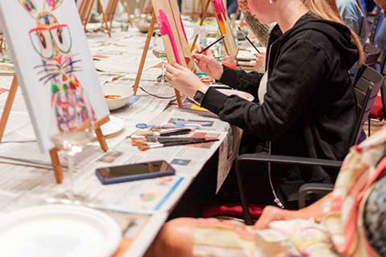  An einem langen Tisch sitzen Frauen mit ihren Staffeleien. 