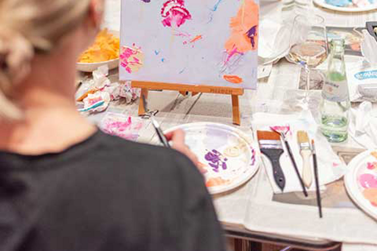  An einem langen Tisch sitzen Frauen mit ihren Staffeleien. 