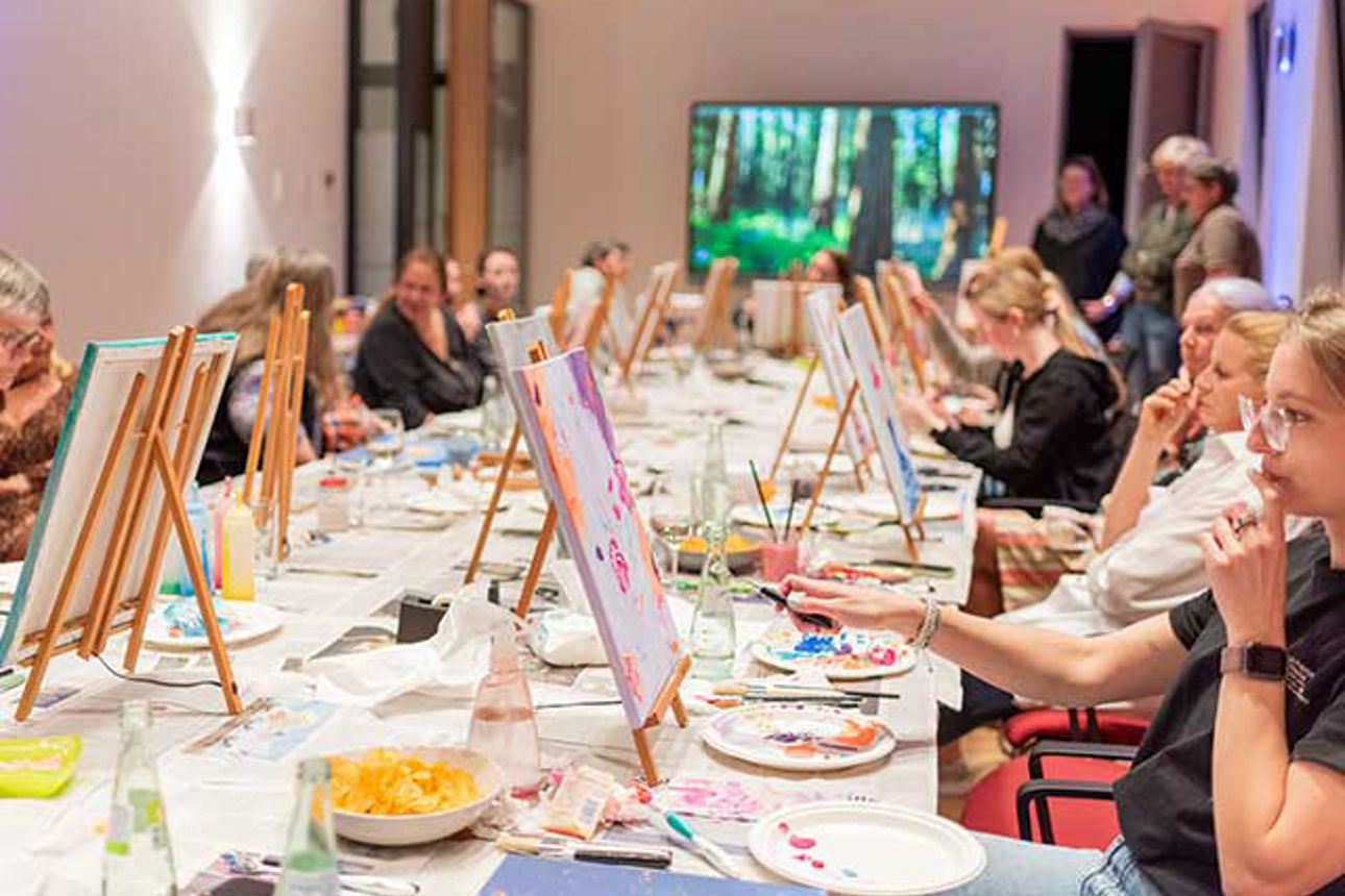  An einem langen Tisch sitzen Frauen mit ihren Staffeleien. 