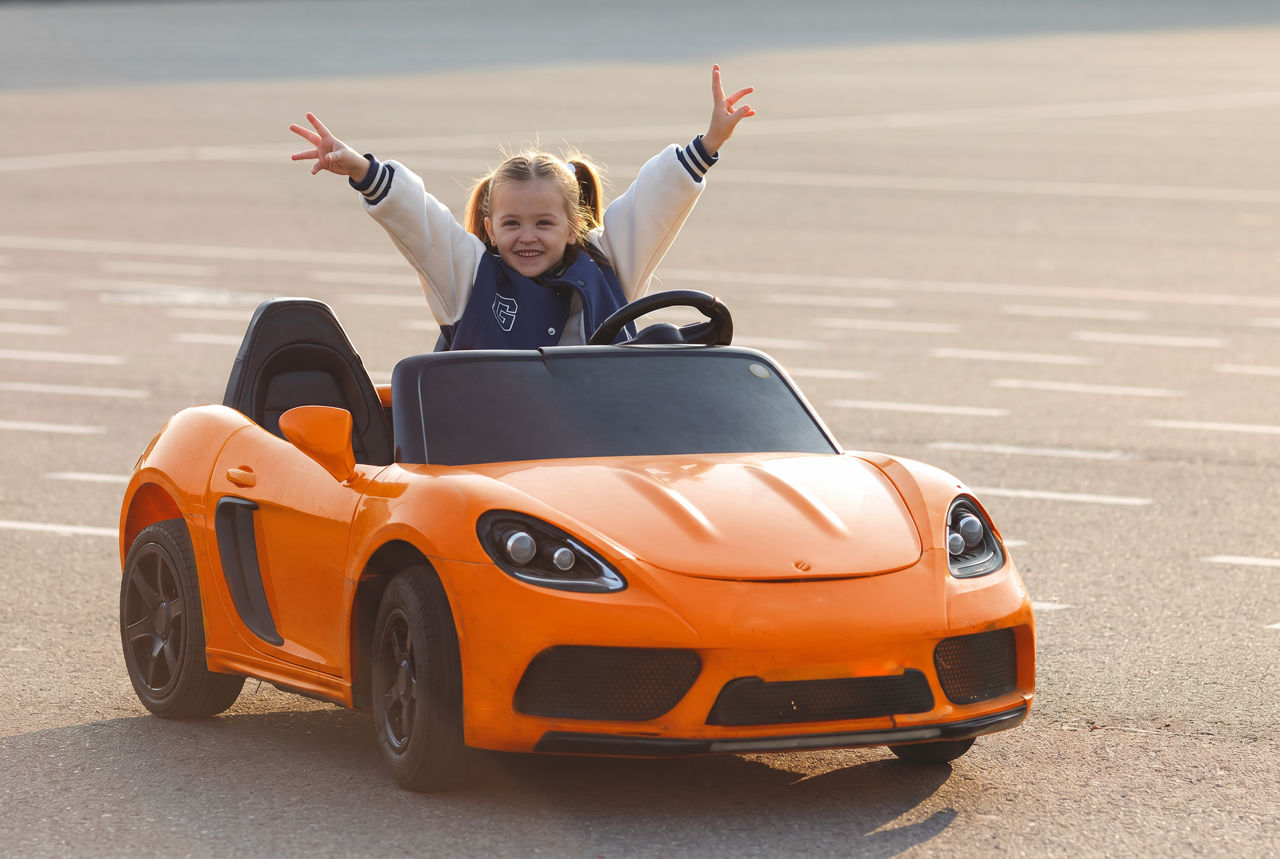 Ein glückliches Mädchen sitzt in einem orangefarbenen Spielzeugauto und streckt beide Arme nach oben.