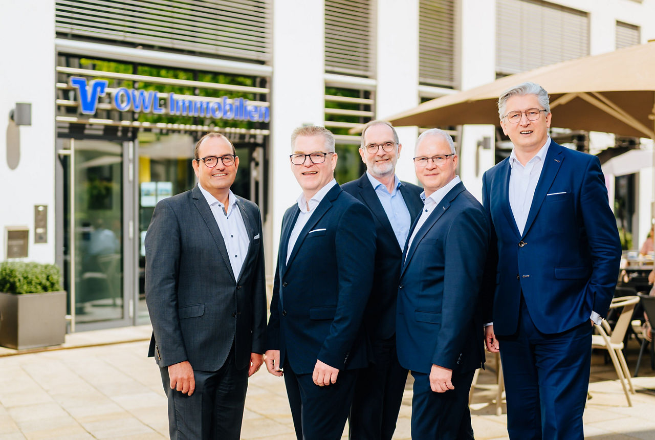 Die drei Geschäftsführer der OWL Immobilien GmbH und zwei Mitglieder des Vorstands der VerbundVolksbank OWl eG stehen vor dem Eingang der OWL Immobilien GmbH in Paderborn.