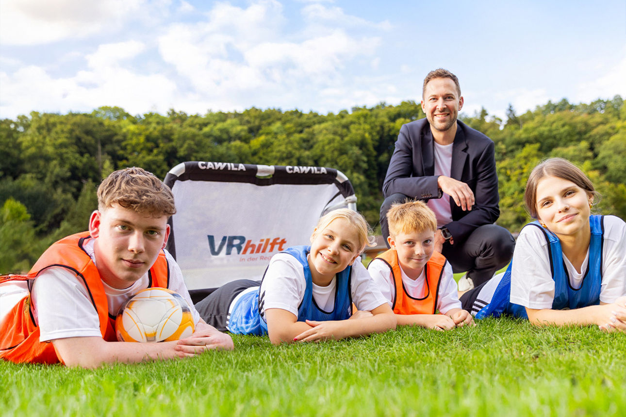 4 Kinder mit Fußballleibchen liegen vor einem Fußballtor auf einer Wiese, im Hintergrund kniet ein Mann in Anzug