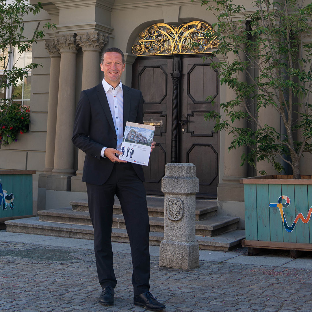 Immobilienberater Patrick Drumm vor einem alten Gebäude im Anzug mit einer Broschüre in der Hand.