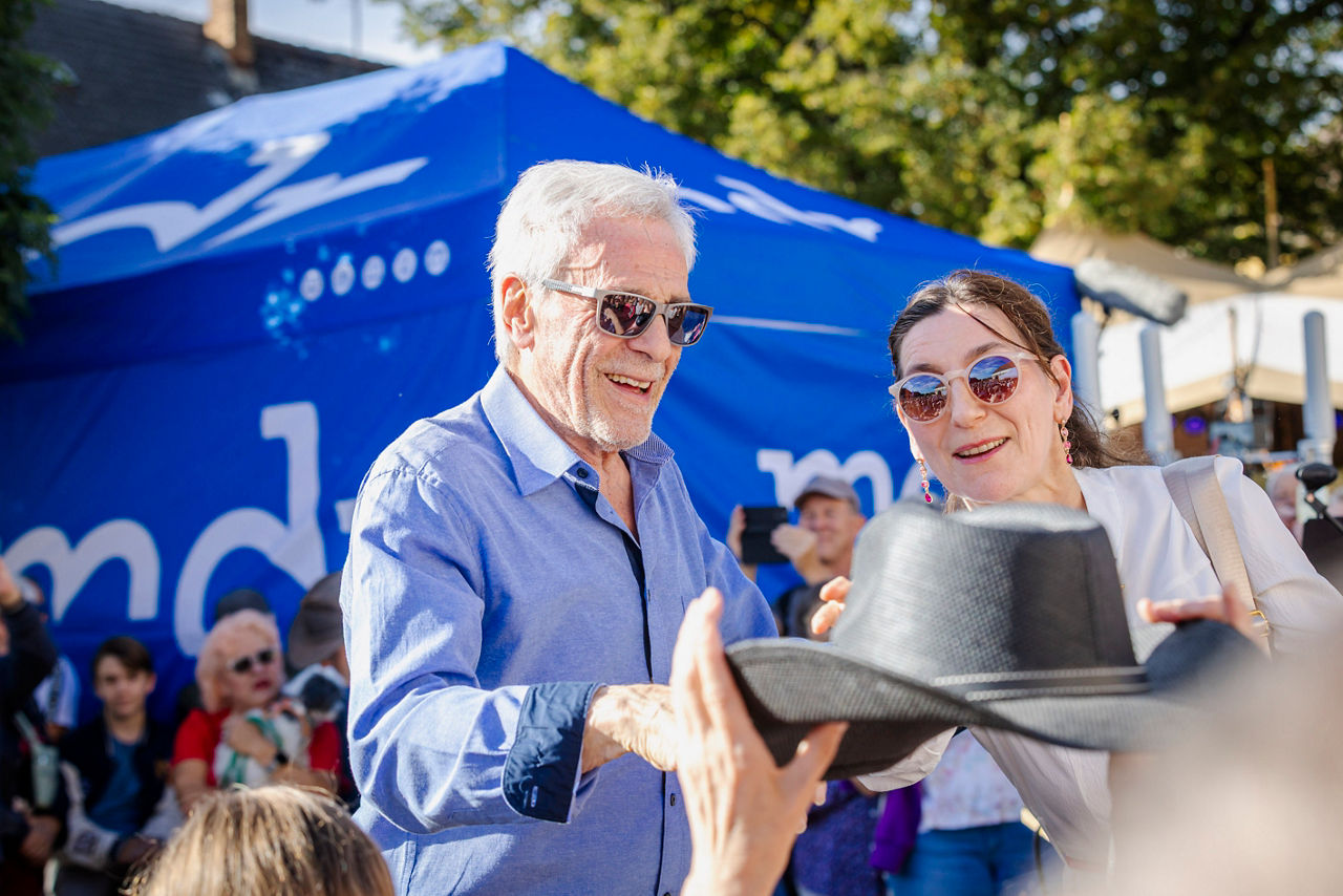im Foto: Filmlegende Gojko Mitić mit seiner Freudin JudithFilmlegende Gojko Mitić begeistert in Heeselicht – MDR überträgt große Show liveDEFA-Star kehrt an Originaldrehort zurück – drei Stunden Autogramme – ungewöhnliche Sandstein-Stele erinnert an „Die Söhne der großen Bärin“Heeselicht. Ein Filmstar, drei Stunden Autogramme und ein Fernseher aus Sandstein: Gojko Mitić hat am Sonnabend die Kirmes in Heeselicht zum Publikumsmagneten gemacht. Der 86-jährige DEFA-Star kehrte an einen Originaldrehort von „Die Söhne der großen Bärin“ zurück. Am Abend wurde der kleine Ort zur großen MDR-Fernsehbühne – inklusive der Einweihung eines ungewöhnlichen Denkmals, das direkt auf die Filmkulisse von einst blickt.Gojko Mitić gehört zu den bekanntesten Gesichtern des DDR-Kinos. Mit seiner Rolle als Häuptling Tokei-ihto in „Die Söhne der großen Bärin“ wurde er in den 1960er-Jahren zum Publikumsliebling. Teile des 1966 erschienenen DEFA-Indianerfilms entstanden in der Sächsischen Schweiz. Heeselicht erinnert bis heute an diese besondere Verbindung zur Filmgeschichte.Am Sonnabend stand Mitić zunächst bei einer Gesprächsrunde im Mittelpunkt. Danach nahm er sich viel Zeit für seine Fans. Rund drei Stunden schrieb der Schauspieler Autogramme, sprach mit Besuchern und posierte für zahlreiche Fotos.Ein Fernseher aus Sandstein zeigt die FilmkulisseBesonders ungewöhnlich ist das neue Denkmal, das am Abend gemeinsam mit Gojko Mitić eingeweiht wurde. Neben einer Bärenfigur steht eine große Stele aus Sandstein – gestaltet wie ein überdimensionaler Fernseher.Wer vor der Stele steht, kann gewissermaßen in den steinernen Fernseher hineinschauen. Durch die Öffnung fällt der Blick direkt in die Landschaft und damit in die Umgebung des ehemaligen Drehortes. Die Natur wird so selbst zum Fernsehbild.Das Denkmal verbindet damit auf ungewöhnliche Weise die regionale Sandsteinlandschaft mit der Filmgeschichte. Besucher sollen nicht nur an die Dreharbeiten erinnert werden, sondern den Bezug zum Originaldrehort unmittelbar erleben können.Bereits im Frühjahr hatte der MDR über das Projekt berichtet. Der Bildhauer Dirk Wachtel arbeitete damals an der ungewöhnlichen Stele. Sie soll gemeinsam mit der Bärenfigur dauerhaft an die Dreharbeiten von „Die Söhne der großen Bärin“ erinnern.Warum ein Bär in Heeselicht stehtAuch die Bärenfigur nimmt Bezug auf den DEFA-Klassiker. Schon der Titel „Die Söhne der großen Bärin“ verweist auf die Geschichte des Films und die Verbindung der Figuren mit der Bärin als symbolischer Stammmutter.Im Mittelpunkt des Films steht der junge Häuptling Tokei-ihto, gespielt von Gojko Mitić. Die Geschichte erzählt von seinem Kampf gegen die Vertreibung seines Volkes aus seiner Heimat.Mit der Bärenfigur und der Sandstein-Stele bekommt Heeselicht damit einen sichtbaren Erinnerungsort an den Film, der den Ort vor rund 60 Jahren mit der DEFA-Filmgeschichte verbunden hat.Unterstützt wurde das Projekt auch von der Volksbank Pirna. Die Bank beteiligte sich über ihre Stiftung an dem Vorhaben. Die Stiftung unterstützt unter anderem Projekte aus den Bereichen Kunst und Kultur sowie Heimatpflege und Heimatkunde in der Region.Heeselicht wird zur großen MDR-BühneAm Sonnabend ging es in Heeselicht aber nicht nur um Filmgeschichte. Die traditionelle Kirmes wurde gleichzeitig zur großen Fernsehproduktion. Der MDR übertrug die Sendung „Hol dir deine Show!“ ab 20.15 Uhr live aus dem Ort.Bereits am Vormittag hatte der Radeberger Spielmannszug das Fest eröffnet. Am Nachmittag sorgte das Duo „Two of Us“ für Livemusik. Nach der Gesprächsrunde mit Gojko Mitić und seiner anschließenden Autogrammstunde ging das Programm mit Richard Säuberlich aus Langenwolmsdorf und Olaf Hobriant aus Warnemünde weiter.Vor Beginn der Liveshow stimmte ein MDR-Warm-up die Besucher auf den Fernsehabend ein. Auf der Bühne standen unter anderem Frank Schöbel, die Band More Than Words und DJ Herzbeat. Auch die Einweihung der Bärenfigur und der Sandstein-Stele wurde in das Abendprogramm eingebunden.Filmklassiker zum Abschluss unter freiem HimmelUm 22 Uhr wurde der Kreis schließlich geschlossen: Unter freiem Himmel lief „Die Söhne der großen Bärin“ noch einmal auf der Leinwand.Damit erlebten die Besucher einen Tag ganz im Zeichen der Filmgeschichte: Der Hauptdarsteller des DEFA-Klassikers war persönlich vor Ort, ein neues Denkmal wurde eingeweiht und am Abend konnte der Film, der Heeselicht mit der DEFA-Geschichte verbindet, noch einmal dort angeschaut werden.Neben der MDR-Show und dem Filmprogramm bot die Kirmes den Besuchern eine Tombola, Spielangebote, regionale Händler sowie Speisen und Getränke.Für Heeselicht war es damit ein außergewöhnlicher Festtag: Gojko Mitić kehrte an einen seiner bekannten Drehorte zurück, schrieb drei Stunden lang Autogramme, der MDR sendete live aus dem Ort – und ein Fernseher aus Sandstein erinnert künftig daran, wo vor rund 60 Jahren ein Stück Filmgeschichte entstand.Foto: Marko Förster26.09.2026 © Marko FörsterWICHTIGE HINWEISE: Jeder Abdruck ist honorar- und umsatzsteuerpflichtig (7 % Ust.)! Die Weitergabe des Bildmaterials an Dritte ist verboten!