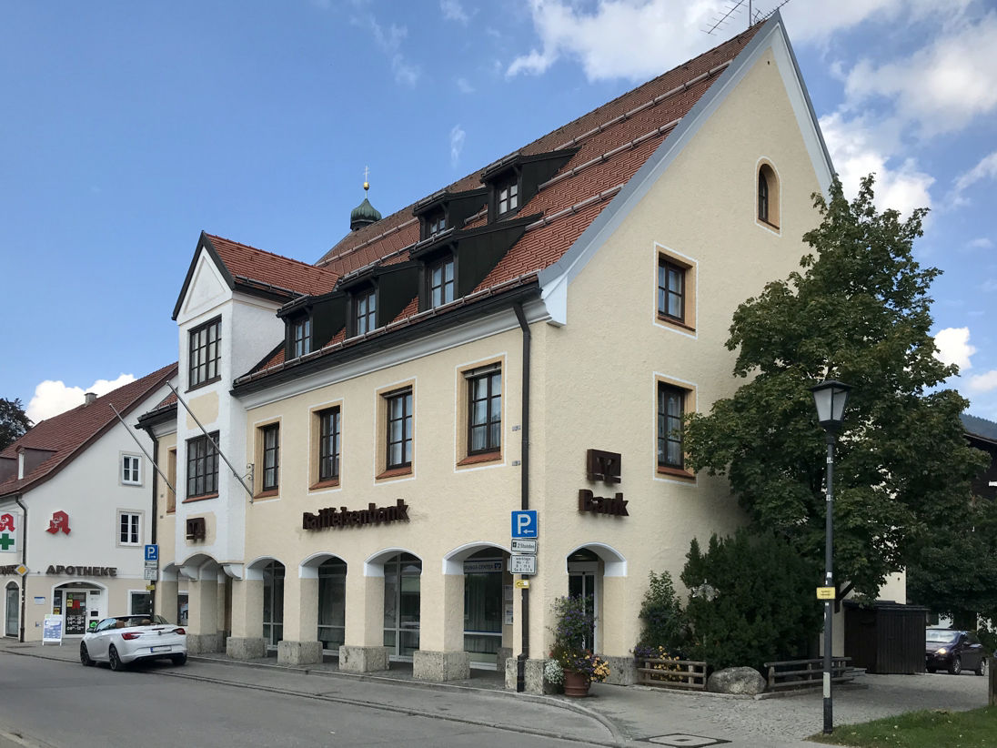 Außenansicht der Raiffeisenbank-Filiale in Lenggries, einem mehrstöckigen Gebäude mit heller Fassade und roten Dächern, mit einem Apotheken-Schild und einem geparkten weißen Auto davor.