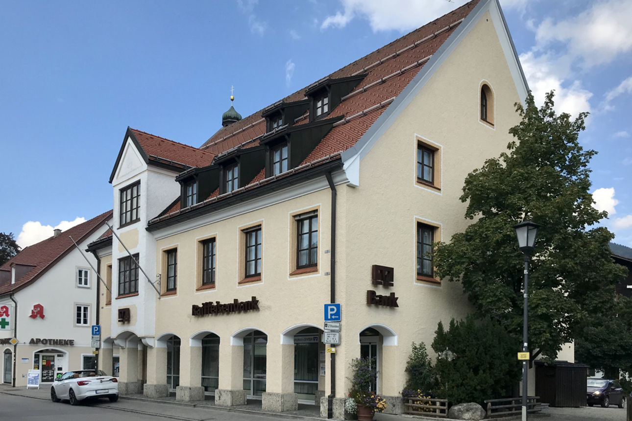 Außenansicht der Raiffeisenbank-Filiale in Lenggries, einem mehrstöckigen Gebäude mit heller Fassade und roten Dächern, mit einem Apotheken-Schild und einem geparkten weißen Auto davor.
