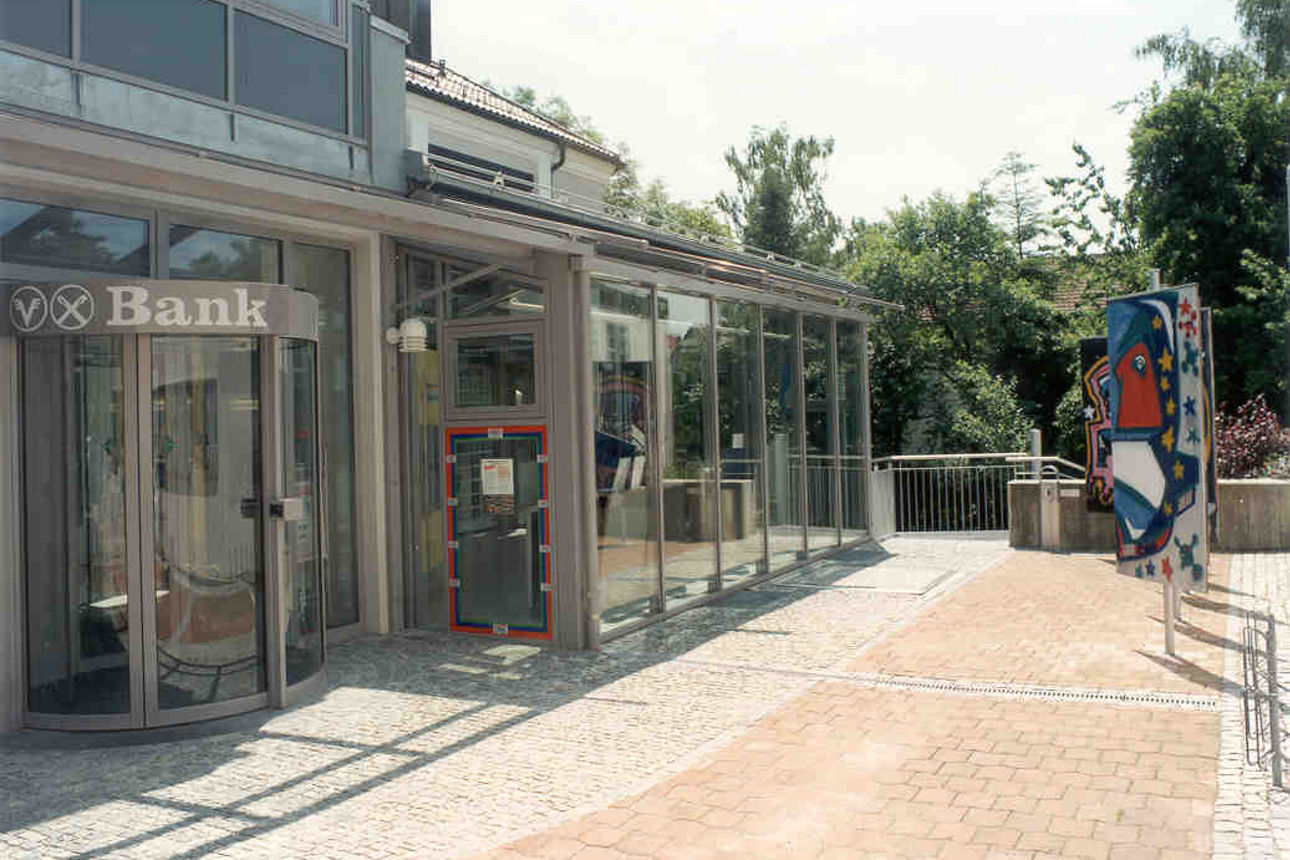 Ein graues Glasgebäude umgeben von Pflanzen und einem Fußgängerweg