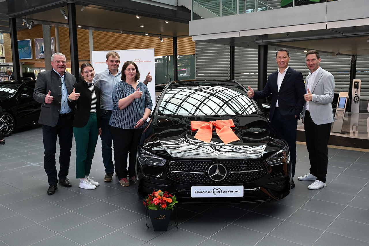 Eine Gruppe von sechs Personen steht in einem Autohaus und lächelt und streckt den Daumen nach oben vor einem nagelneuen schw