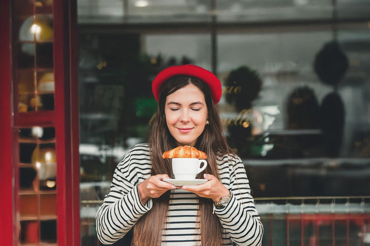 Junge Frau mit einer Baskenmütze riecht mit geschlossenen Augen an einem Croissant, das auf einer Tasse Kaffee liegt.