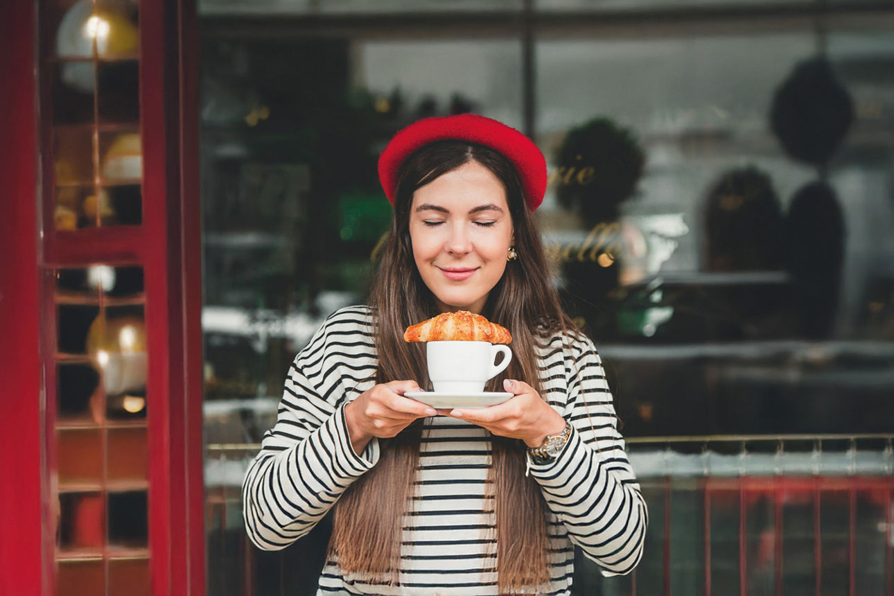 Frau mit Crossaint auf einer Tasse. Sie trägt ein gestreiftes Oberteil und rotes Barrett
