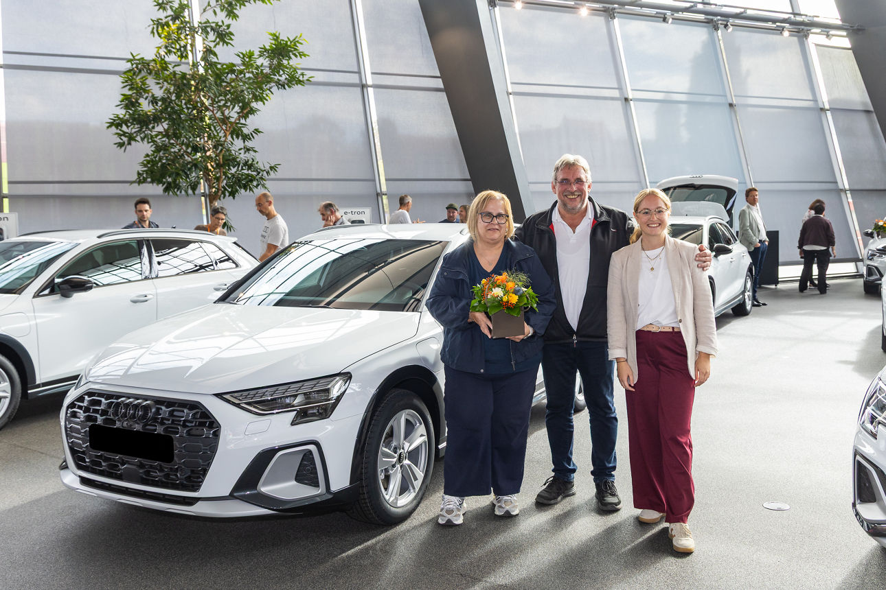 Drei lächelnde Personen stehen neben einem weißen Audi in einem hellen Autohaus.