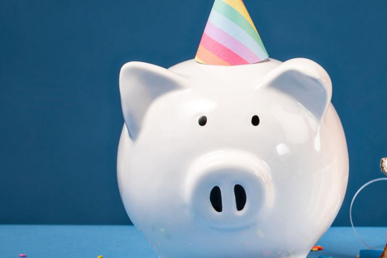 Ein Sparschwein mit Parthütchen steht neben einem Stück Geburtstagskuchen mit einer Kerze