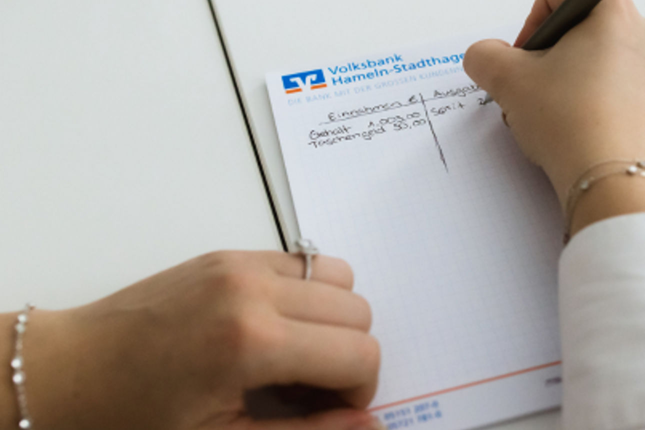 Eine Person schreibt auf einem Notizblock der Volksbank Hameln-Stadthagen handschriftliche Notizen an einem Schreibtisch.