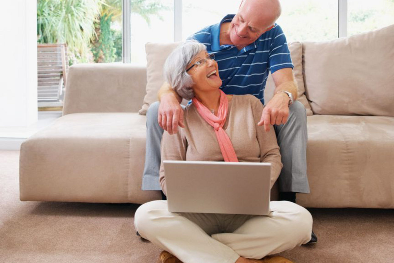 Älteres Paar sitzt lächelnd auf dem Boden vor einem Sofa und schaut gemeinsam auf einen Laptop in einem hellen Wohnzimmer.