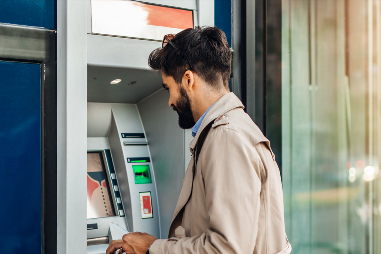 Junger Mann mi dunklen Haaren in hellem Parker vor Geldautomat