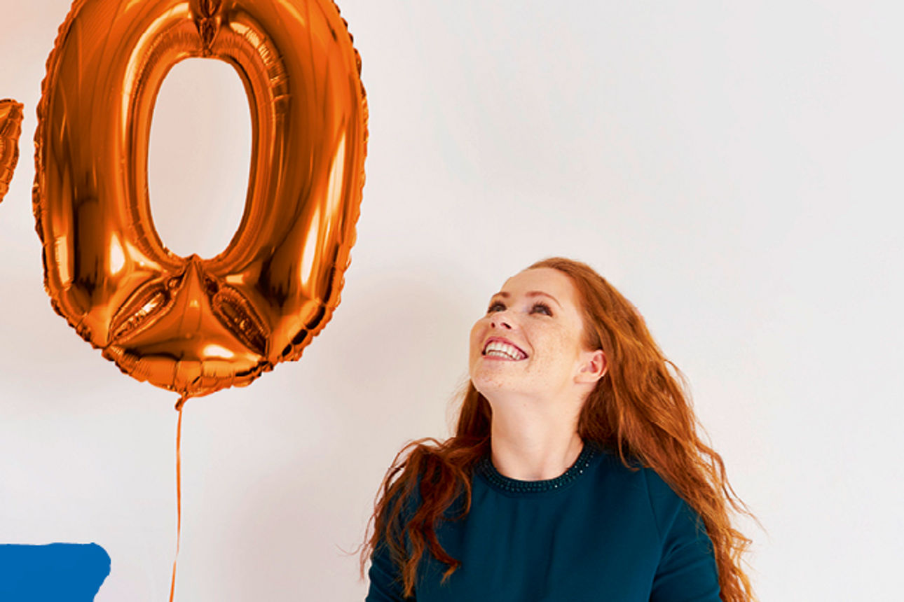 Eine Frau steht neben 2 Luftballons mit den Ziffern 2 und 0