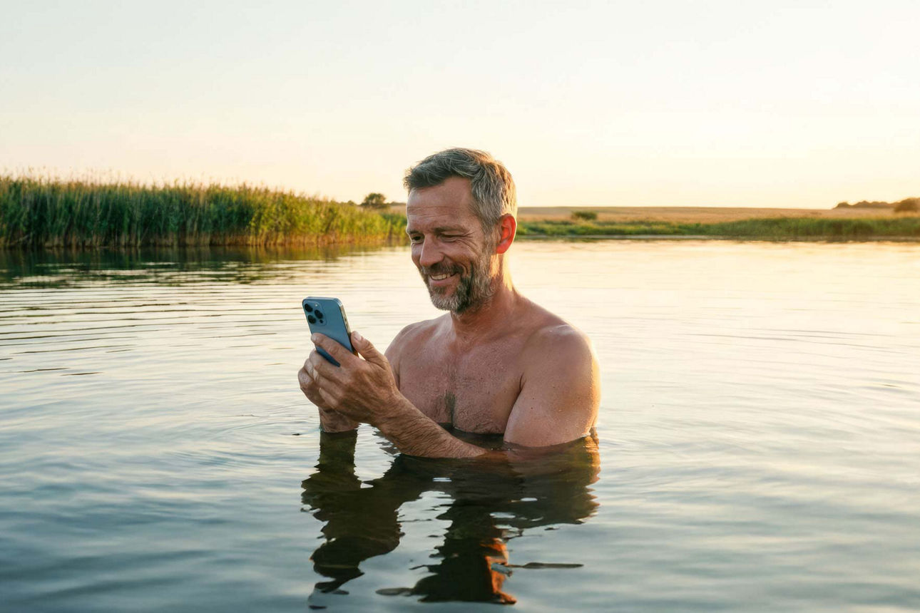 Ein Mann badet in einem See und hat sein Handy in der Hand (KI generiert)