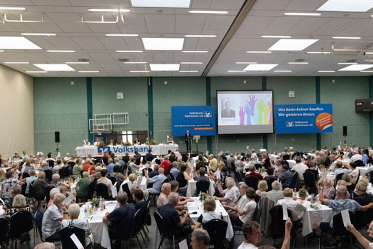 Eine große Gruppe von Menschen sitzt an Tischen in einem turnhallenähnlichen Raum und nimmt an einer Präsentation teil. 