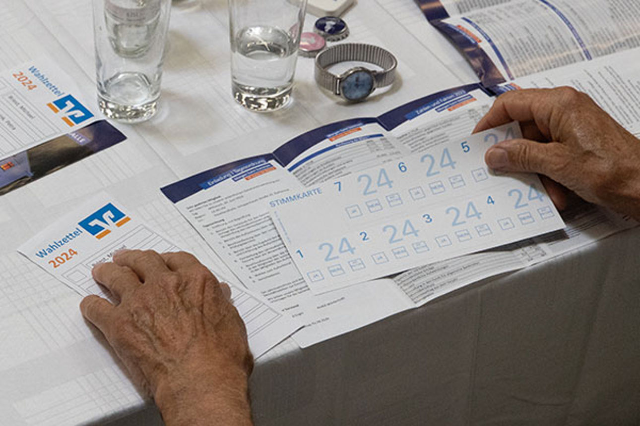 Eine Person füllt an einem Tisch mit einem Glas Wasser, einer Uhr und anderen Stimmzetteln einen Stimmzettel aus, auf dem die