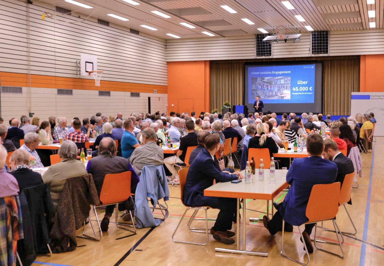 Eine große Bankversammlung mit vielen Kunden in einer Turnhalle und einer Bühne.