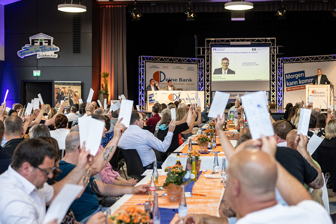 Teilnehmende stimmen bei einer Generalversammlung per Handzeichen in großer Halle ab.