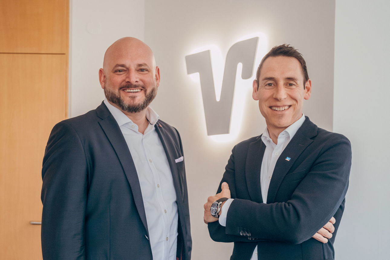 Zwei Männer lachend vor dem Volks-/Raiffeisenbank Logo im Hintergrund