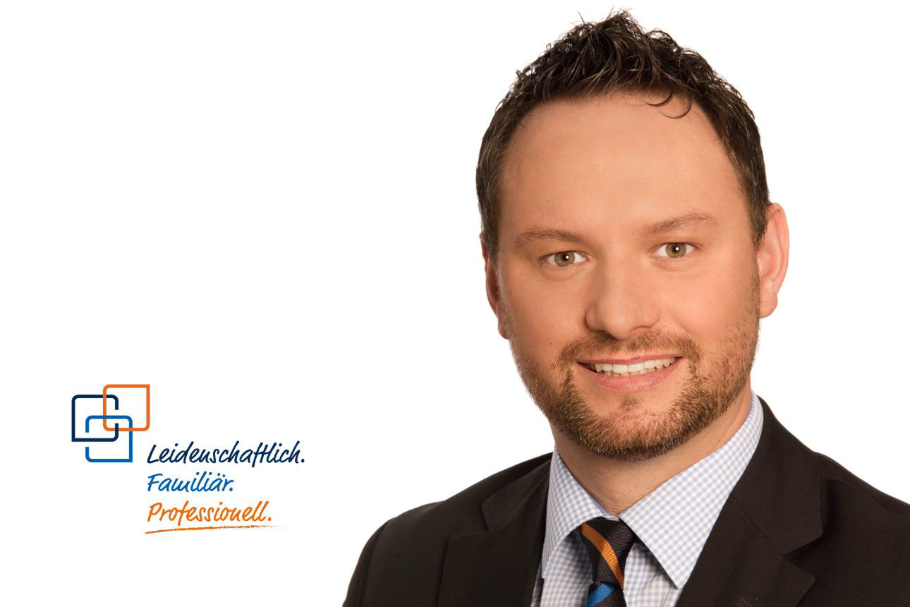 Porträt von Georg Rausch, einem lächelnden Mann mit kurzen dunklen Haaren und Bart im dunklen Anzug mit kariertem Hemd und gestreifter Krawatte. Daneben das Firmenlogo und der Slogan "Leidenschaftlich. Familiär. Professionell."