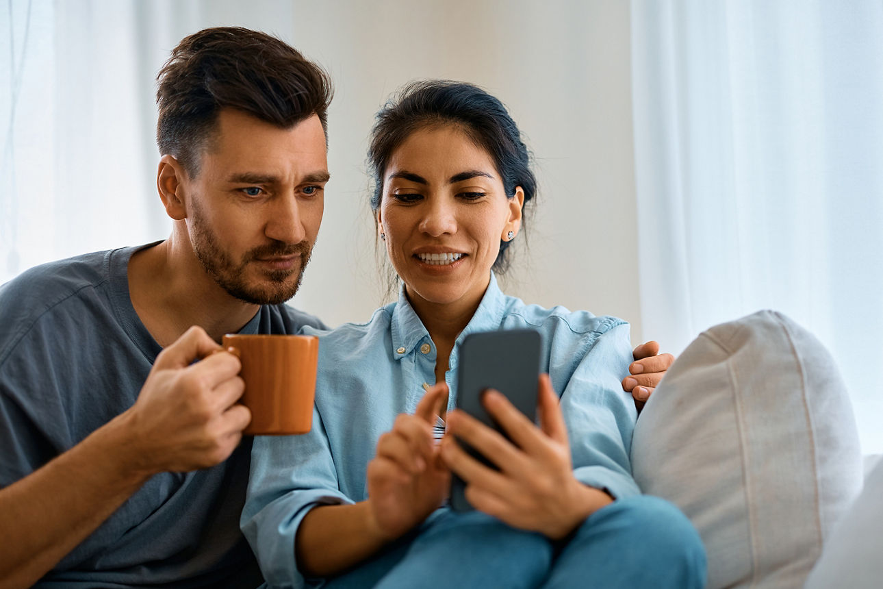 Zwei Personen sitzen auf einem Sofa, eine hält eine Tasse, die andere zeigt auf ein Smartphone.
