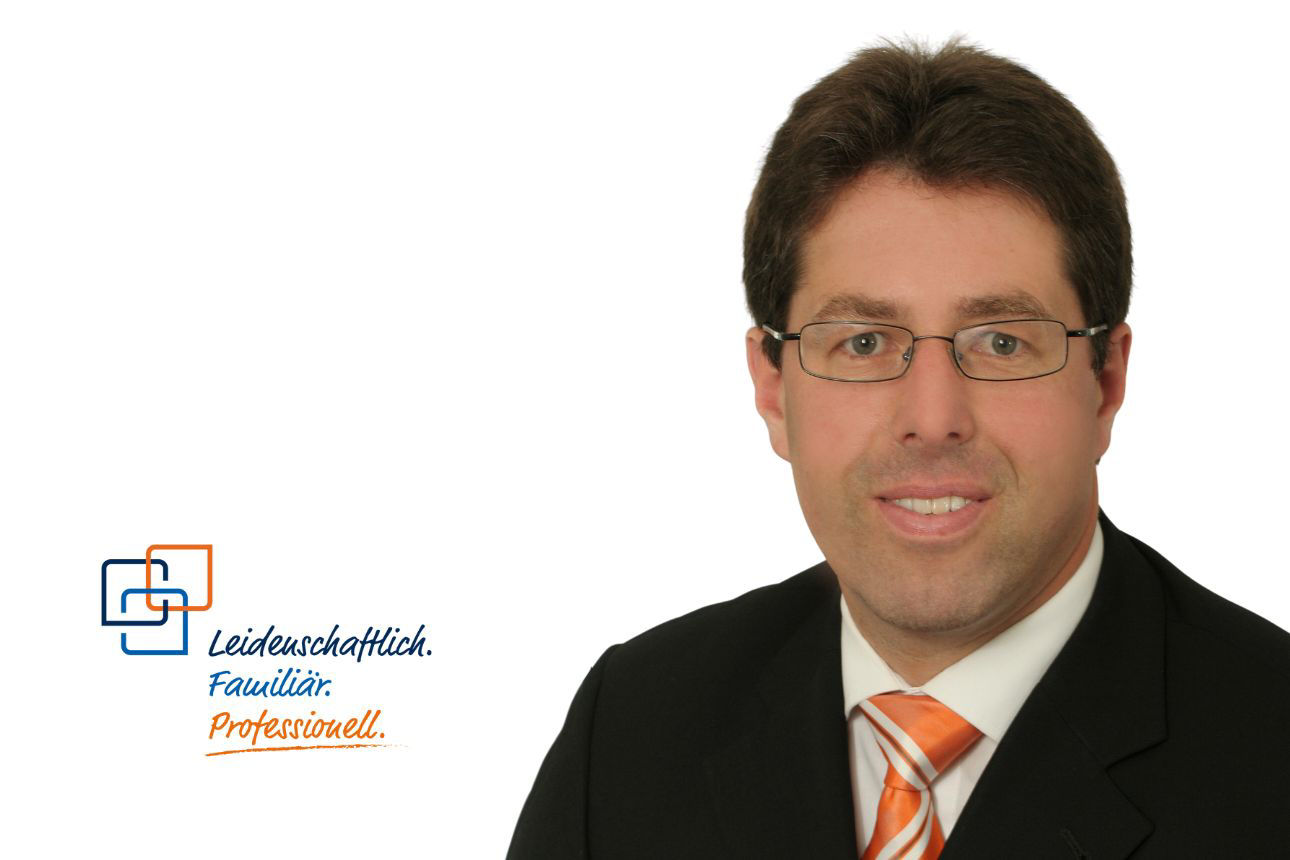 Gerhard Greisinger, ein Mann mit Brille und kurzen, dunklen Haaren, lächelt freundlich und trägt einen dunklen Anzug mit einer gestreiften orange-weißen Krawatte.